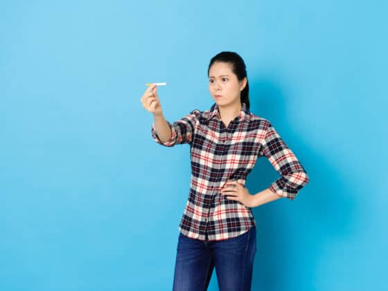 A woman looking confused at her cigarette wondering how long does nicotine stay in your system. Set against blue background.