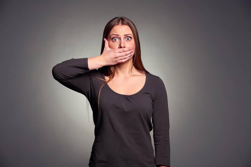 young adult white woman holding hand over her mouth over grey background