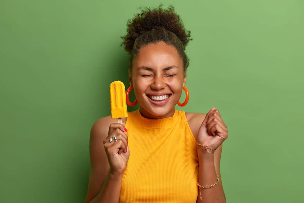 Happy dark skinned millennial girl eats yellow ice cream of mango flavor, clenches fist with anticipation of something awesome happen, has toothy smile, wears bright summer clothes, isolated on green