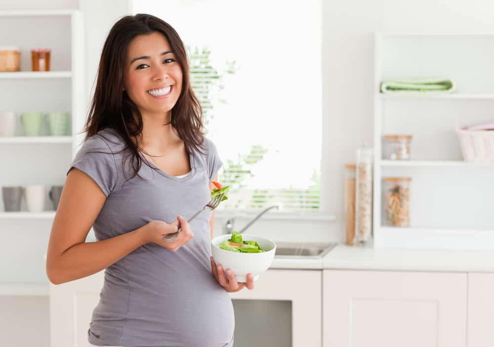 Attractive pregnant woman holding a bowl of salad while standing in the kitchen
